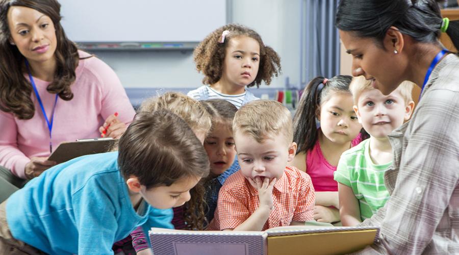 Bild på barn som sitter på golvet och tittar i en bok tillsammans med två pedagoger.